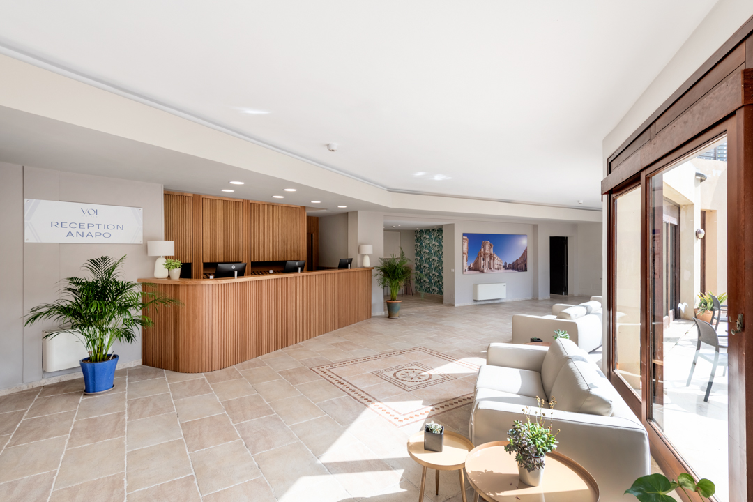 Reception area at VOI Arenella Resort with wooden desk, tile flooring, cream seating, and open doors to a sunlit terrace.