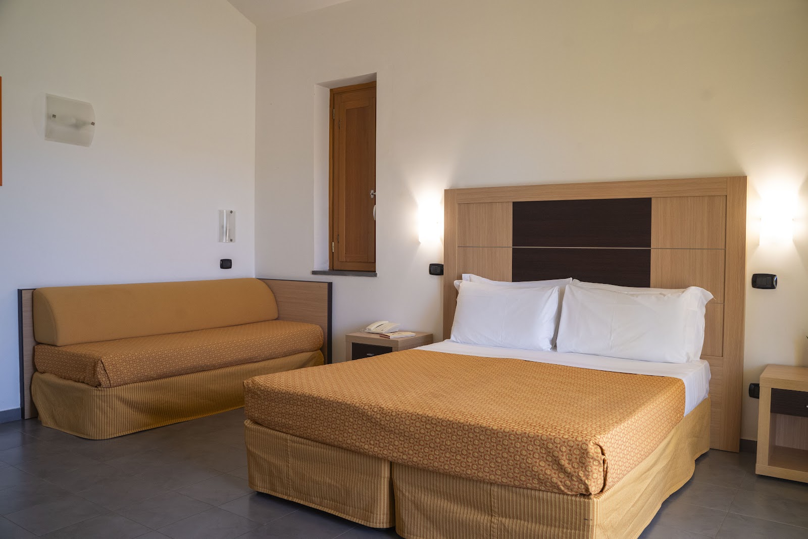 Hotel room with queen bed with white pillows and gold bedcover, matching sofa, wood headboard, and sconces at VOI Baia di Tindari Resort.