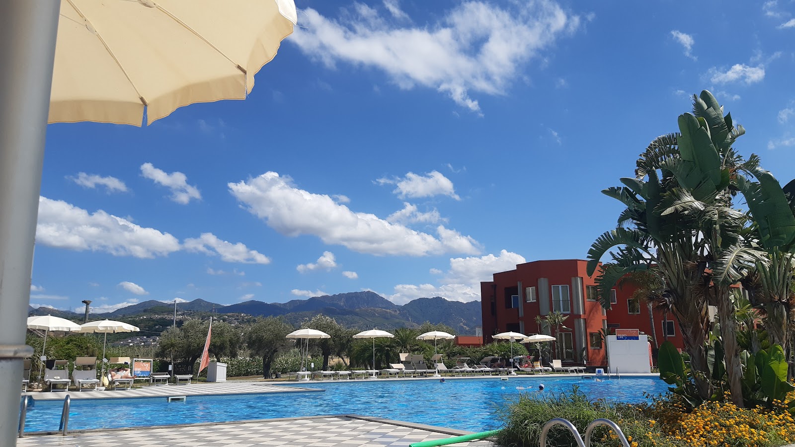 Large blue pool with sun loungers and cream umbrellas lining the deck, terracotta and cream resort buildings behind surrounded by palm trees.