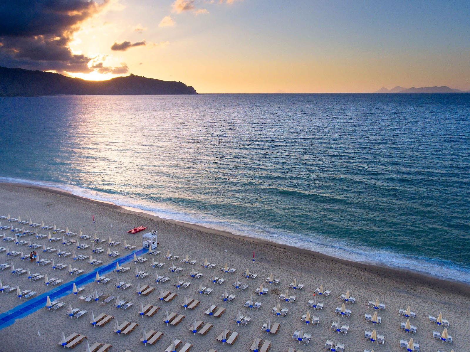 Sunset view over the Tyrrhenian Sea from VOI Baia di Tindari beach, with rows of white loungers and umbrellas lining the sand.