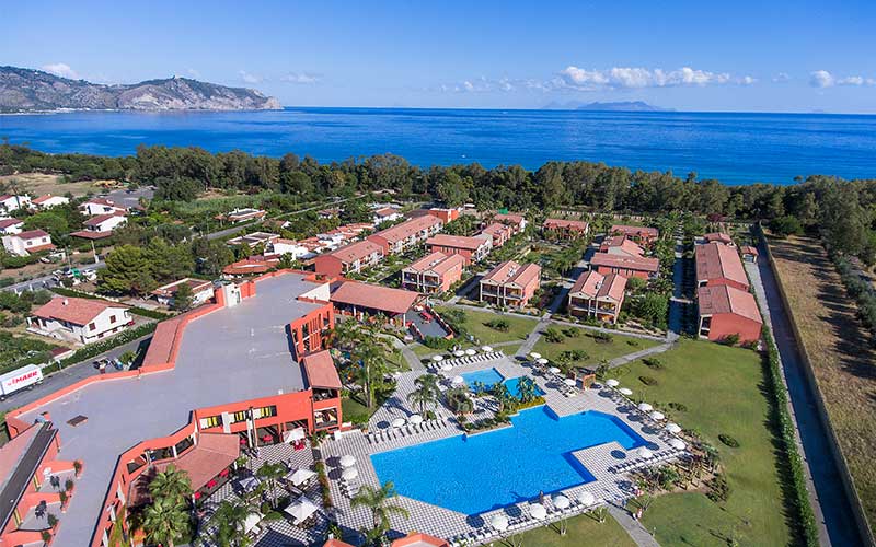 Aerial view of VOI Baia di Tindari Resort with red-roofed buildings, blue pool, and manicured grounds overlooking the Mediterranean Sea and coastal mountains.