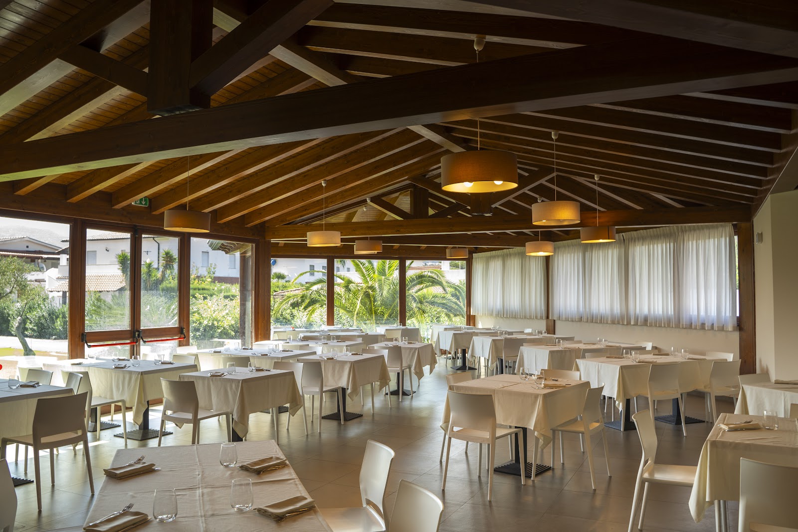 Dining room at VOI Baia di Tindari Resort with exposed wooden beams, pendant lights, tables set for service, and views of tropical gardens through large windows.