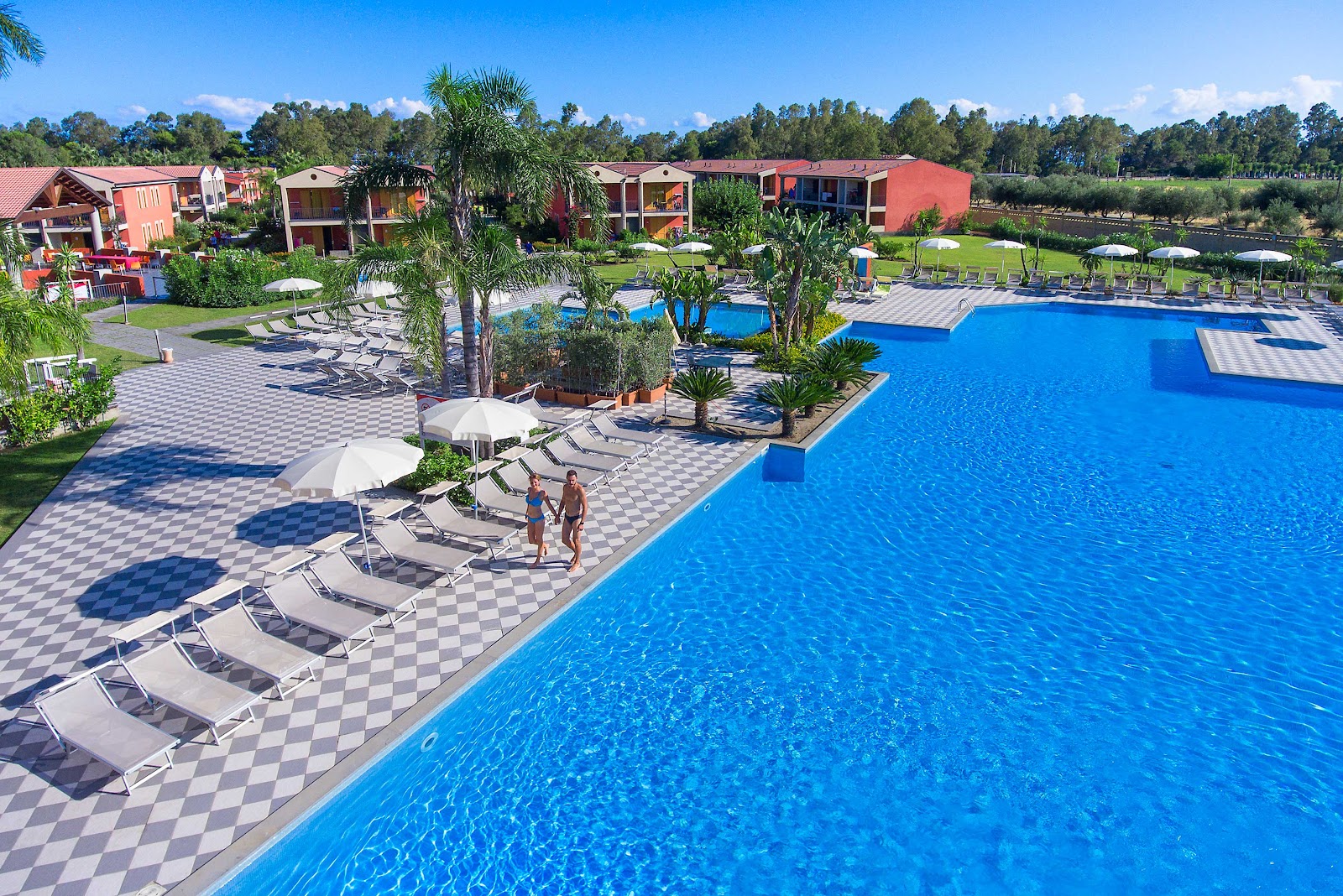 Large blue swimming pool with lounge chairs and white umbrellas, red resort buildings, and palm trees at VOI Baia di Tindari Resort.