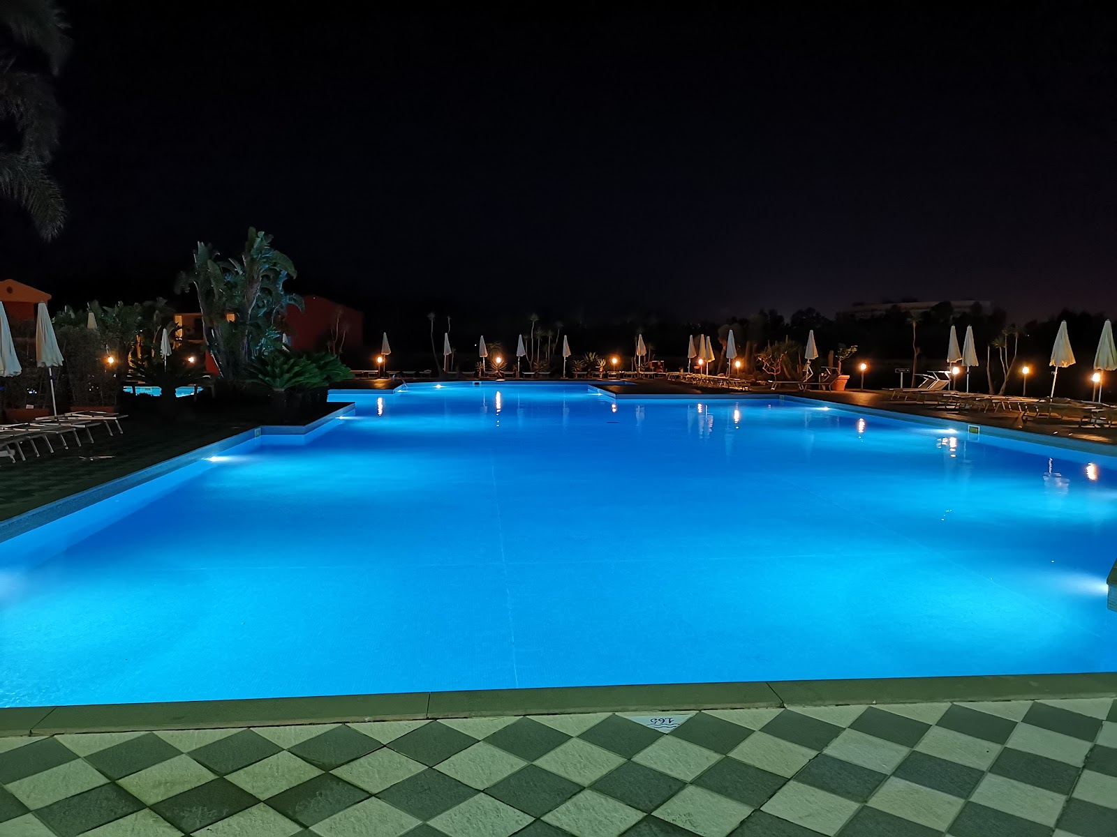 Illuminated pool at night with checkered tile deck, white umbrellas, lounge chairs, and palm trees lining the resort grounds.