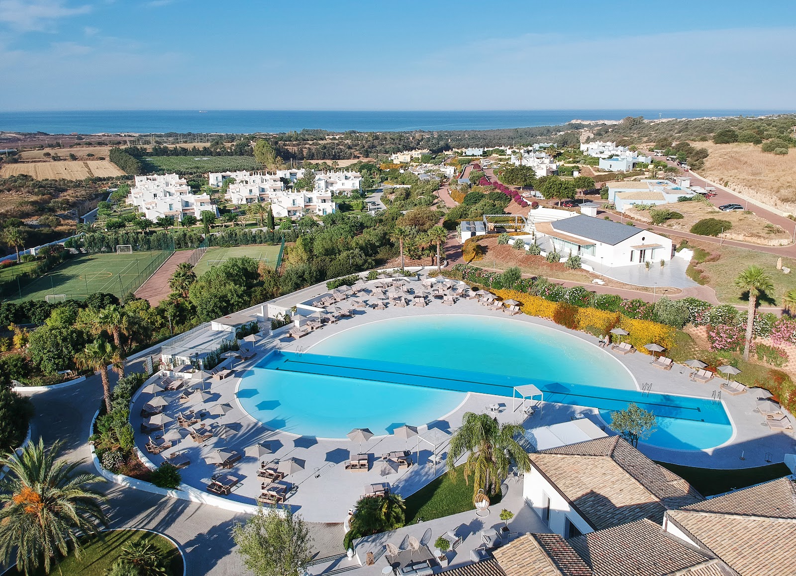 Resort pool with turquoise water and lounge chairs surrounded by white buildings, green grounds, and Mediterranean coastline in the distance.
