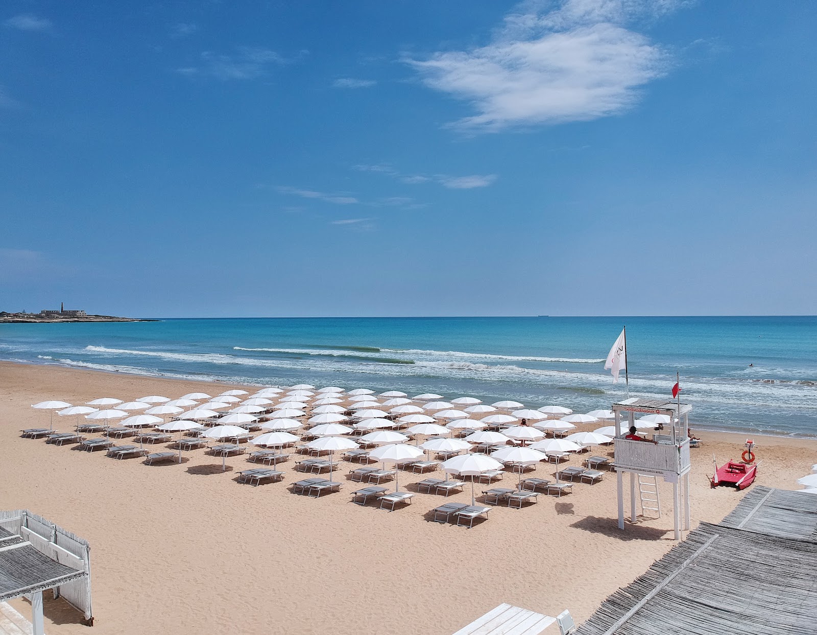 Rows of white umbrellas and lounge chairs line the sandy beach at VOI Marsa Siclà Essentia, with turquoise water and a lifeguard station visible.