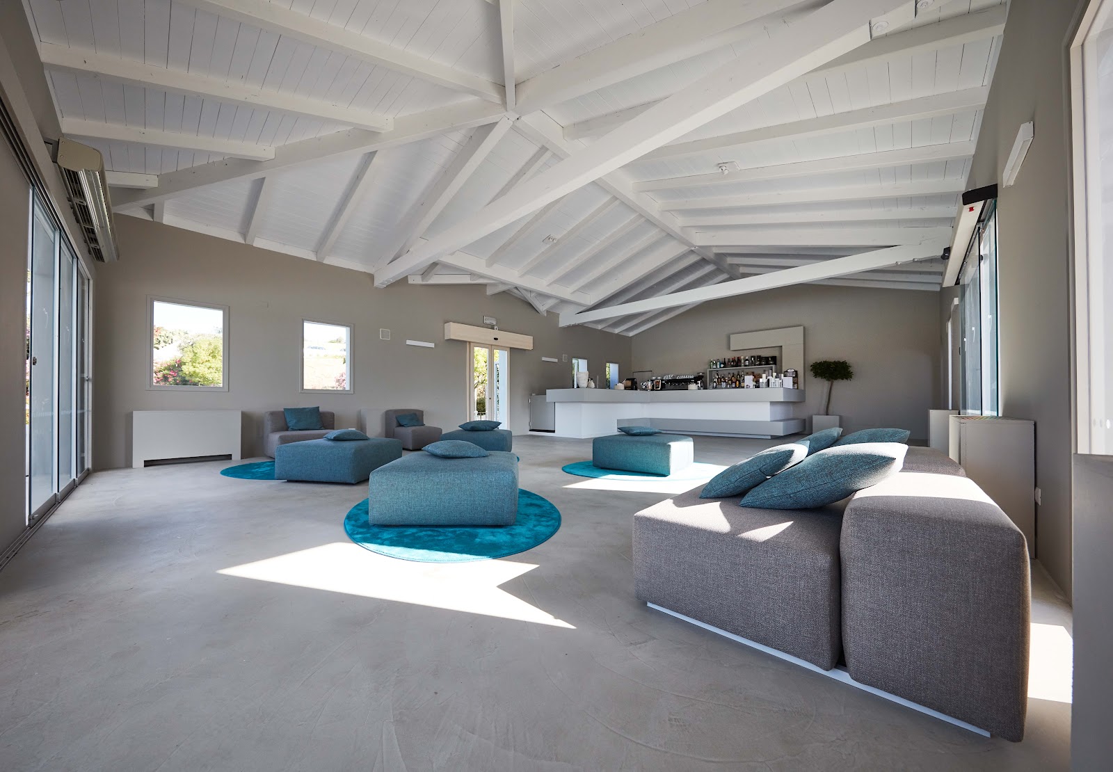 Modern spa lounge with white exposed beam ceiling, gray walls, and blue upholstered seating arranged throughout the polished concrete floor.