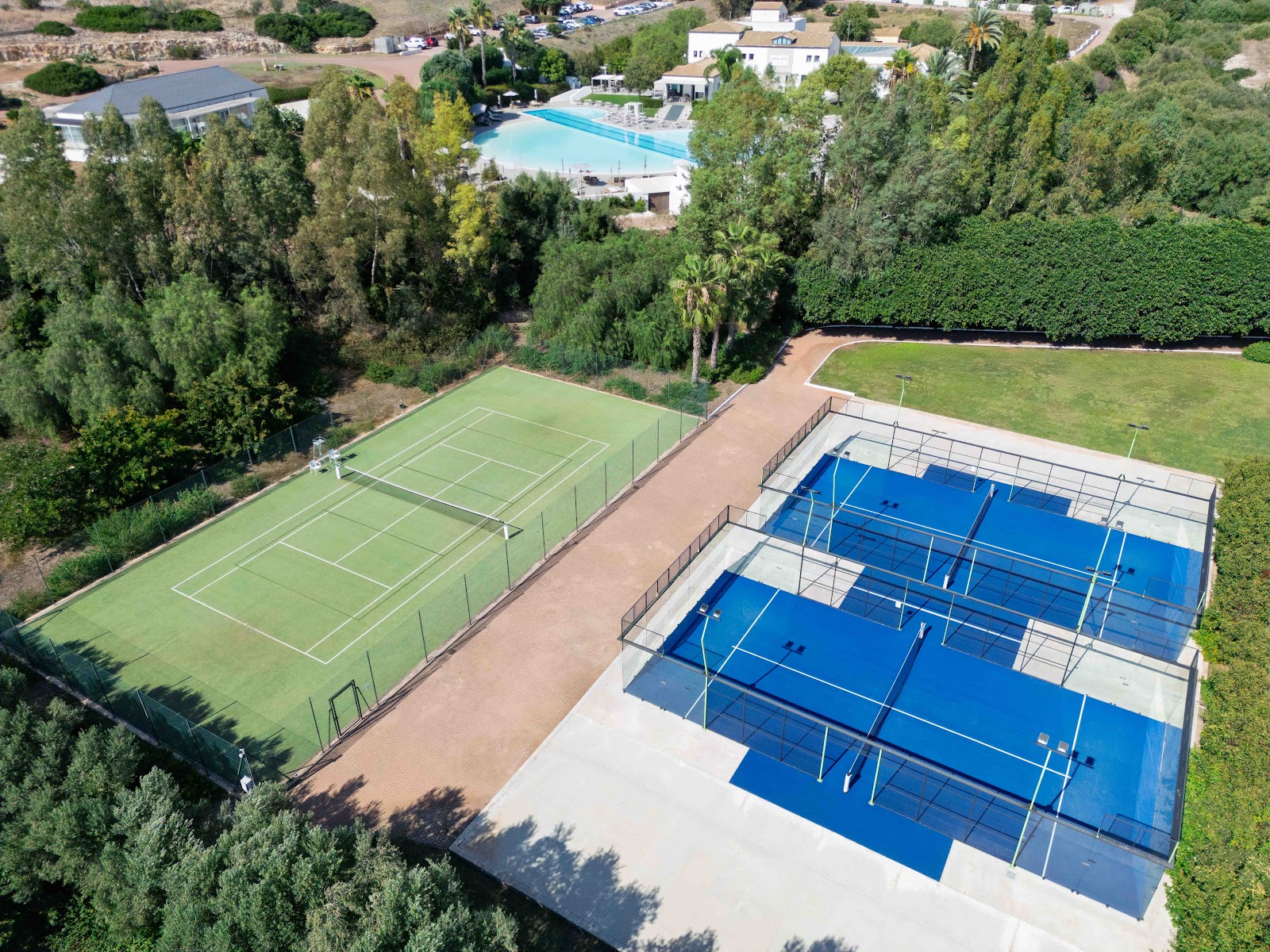 Aerial view of the resort's sports facilities including three blue padel courts, a grass tennis court, and a swimming pool surrounded by pine trees and manicured grounds.