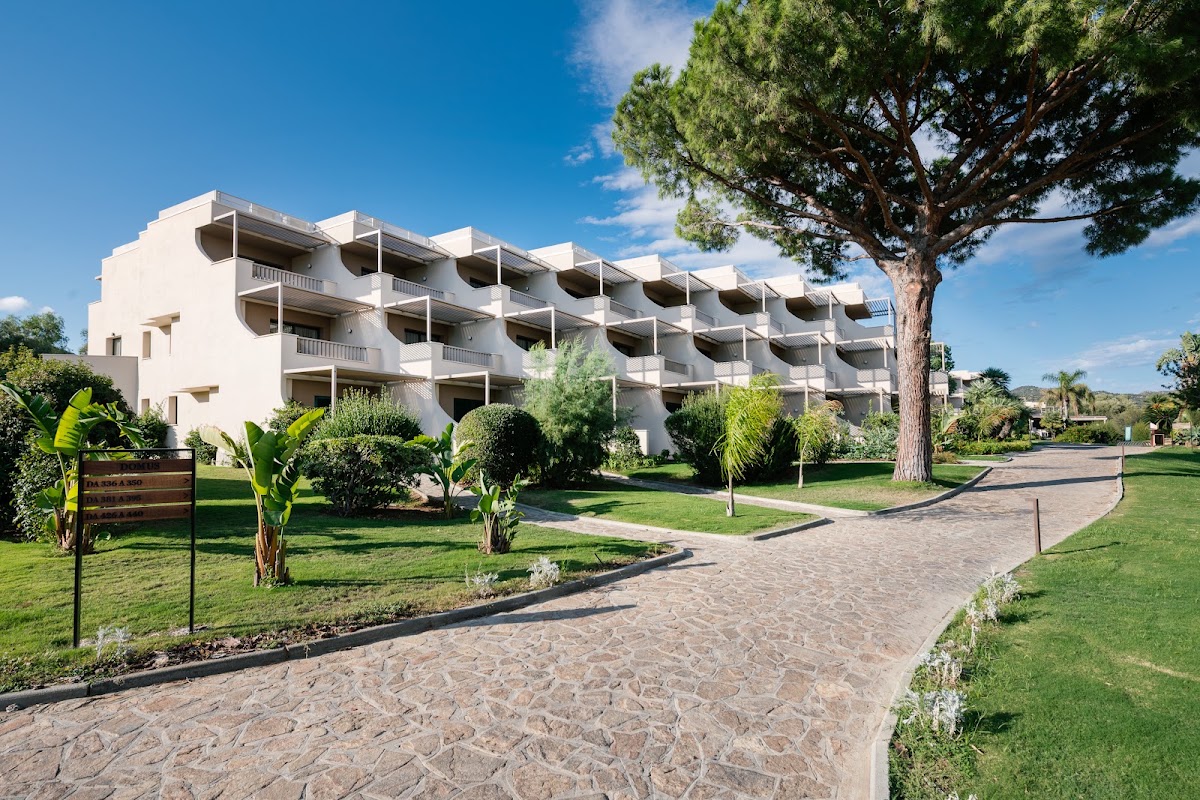 White multi-story resort building with balconies set among tropical landscaping, paved pathway, and manicured lawns under clear blue sky.