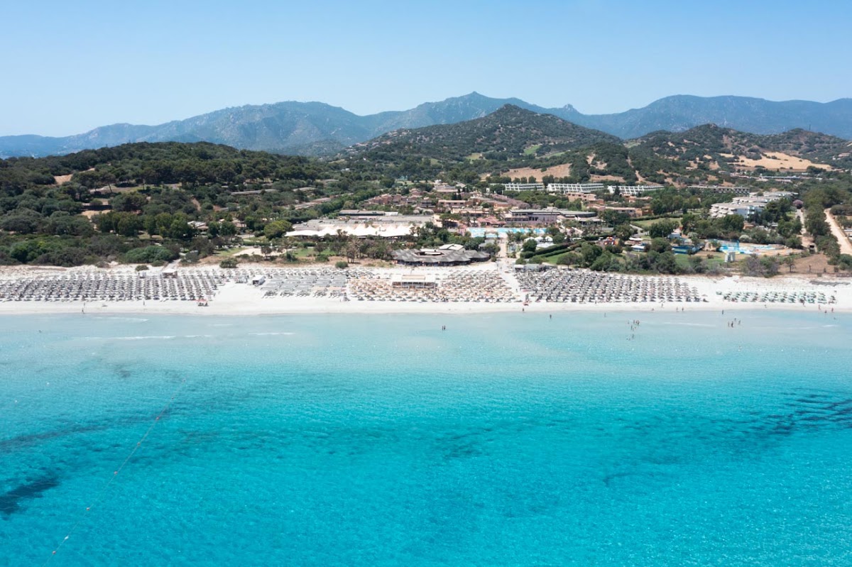 Turquoise Mediterranean waters meet a sandy beach lined with umbrellas and loungers at VOI Tanka Resort, with forested mountains beyond.