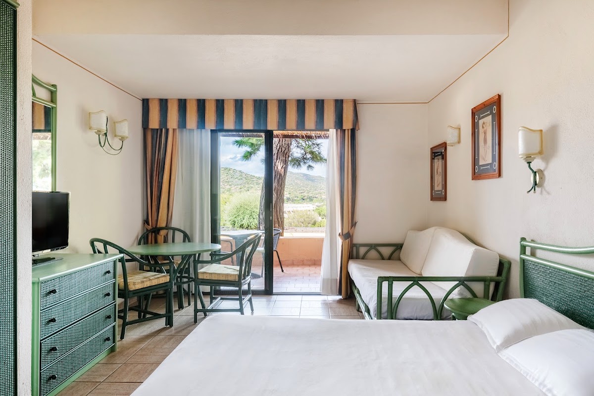 Bright hotel room with two beds with green frames, green dresser, dining table, and terrace overlooking Mediterranean hills and pine trees.