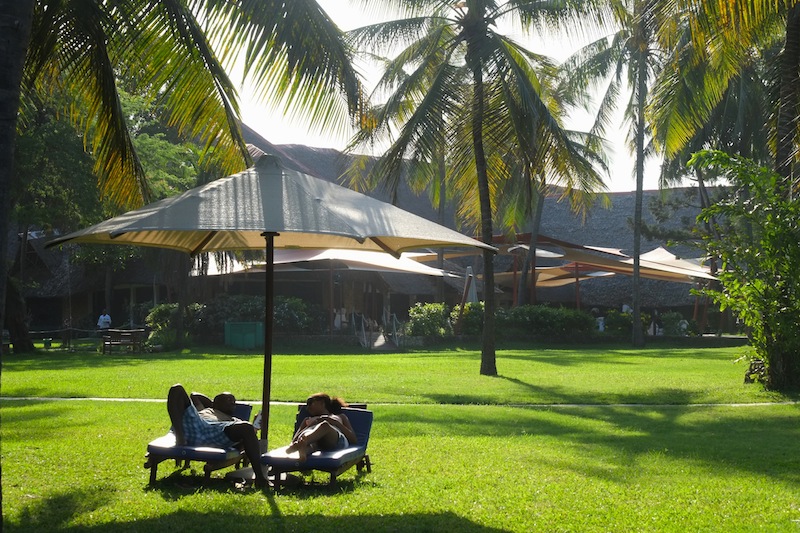 Guests relax on lounge chairs beneath a shade sail on the manicured lawn at Voyager Beach Resort, with palm trees and a stone building in the background.