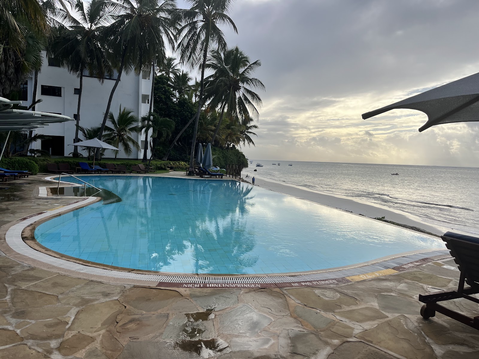 Curved pool with blue water overlooking sandy beach and ocean at Voyager Beach Resort, white building and palm trees behind.