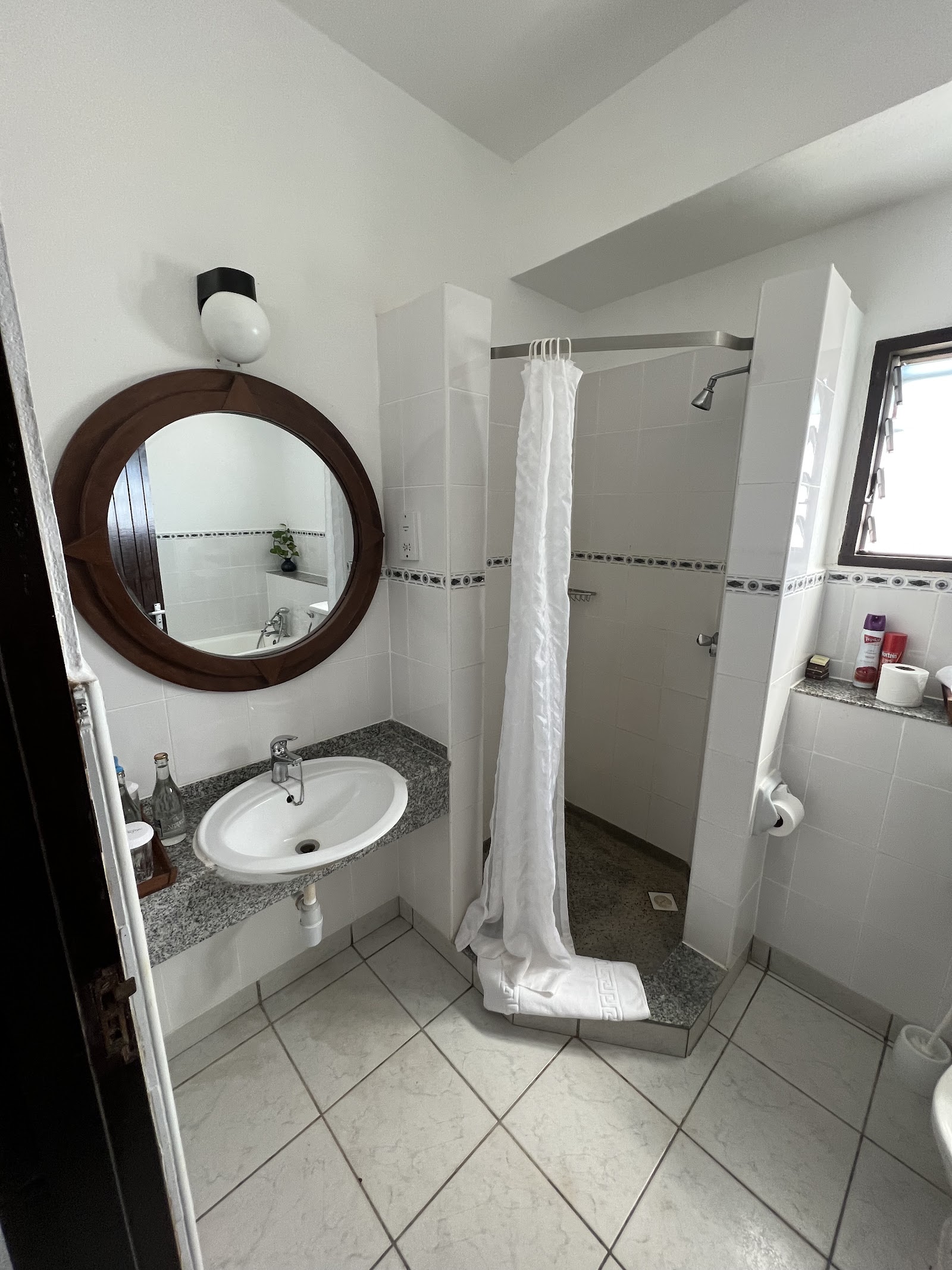 Resort bathroom with white tile walls, round mirror above sink with gray countertop, and walk-in shower with white curtain.