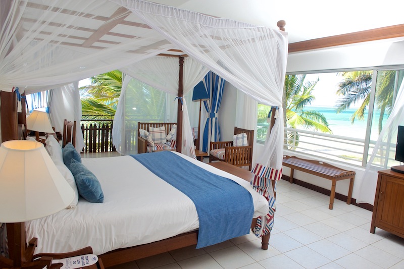 Four-poster bed with white canopy and blue runner in a spacious beach room overlooking palm trees and turquoise ocean at Voyager Beach Resort.