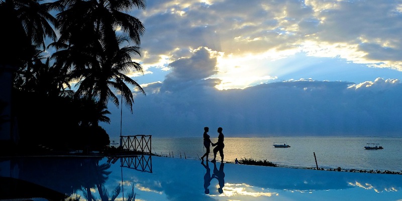 Two guests stand on an infinity pool deck at Voyager Beach Resort with palm trees, calm ocean, and moored boats beneath a dramatic cloudy sky.