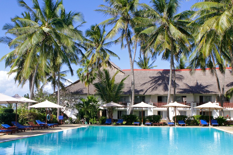 Three guests relax under a shade sail at Voyager Beach Resort with tropical drinks, turquoise ocean and green vegetation visible behind them.
