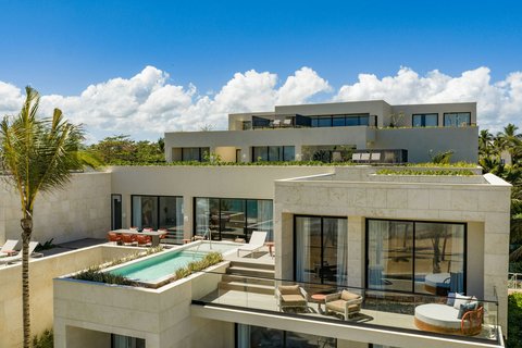 Modern multi-level resort building with cream-colored exterior, rooftop pool, lounge seating, and palm trees under blue sky with white clouds.