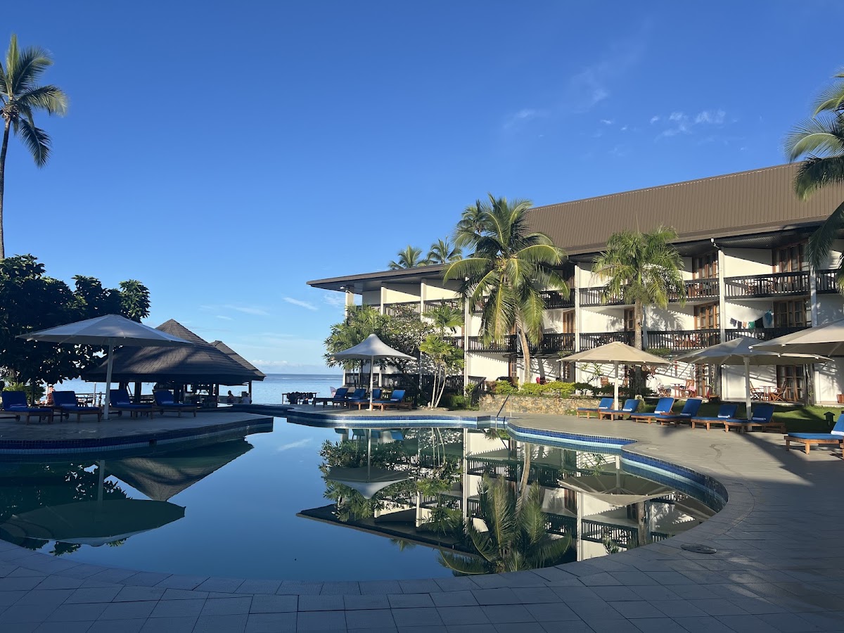 Resort pool reflecting palm trees and multi-story building with covered lounge areas and ocean view in background at Warwick Fiji.