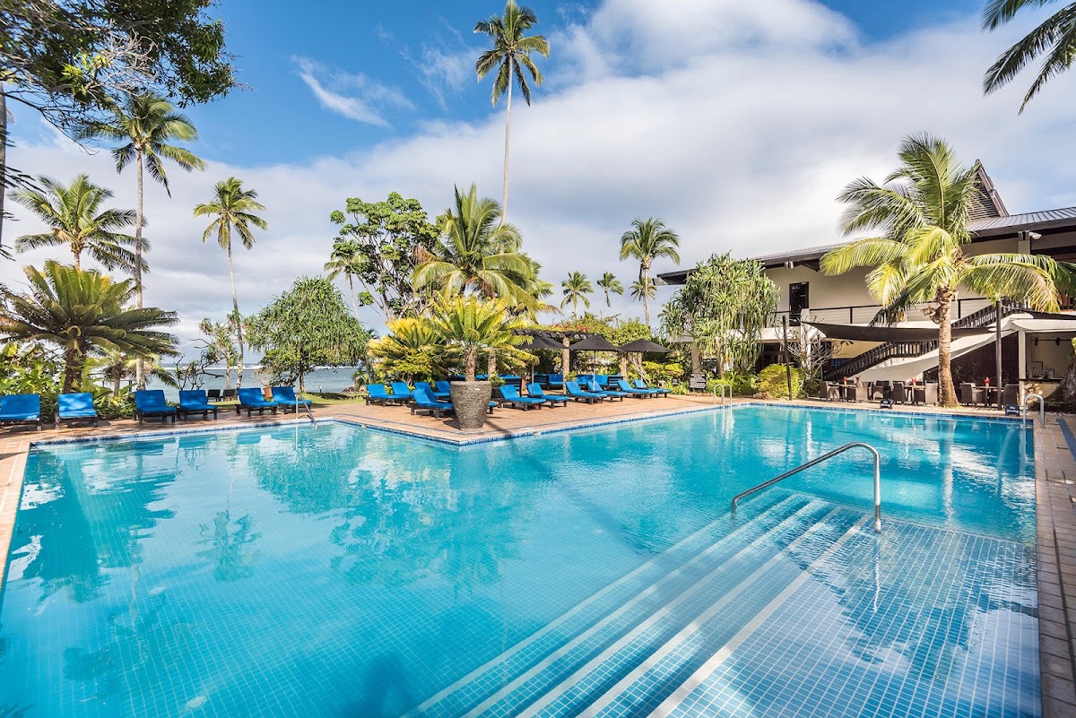 Resort pool with turquoise water, blue loungers, palm trees, and multi-story hotel buildings under partly cloudy sky.