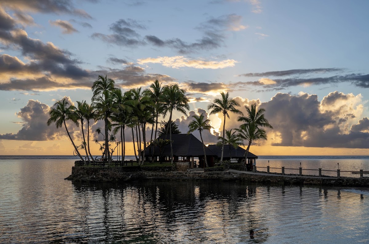 Small island with thatched-roof bungalows and palm trees at sunset, connected by wooden walkway over calm lagoon water.