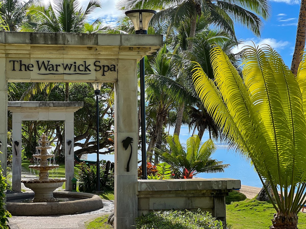 The Warwick Spa entrance framed by a concrete archway with fountain, surrounded by palm trees and tropical vegetation overlooking the ocean.