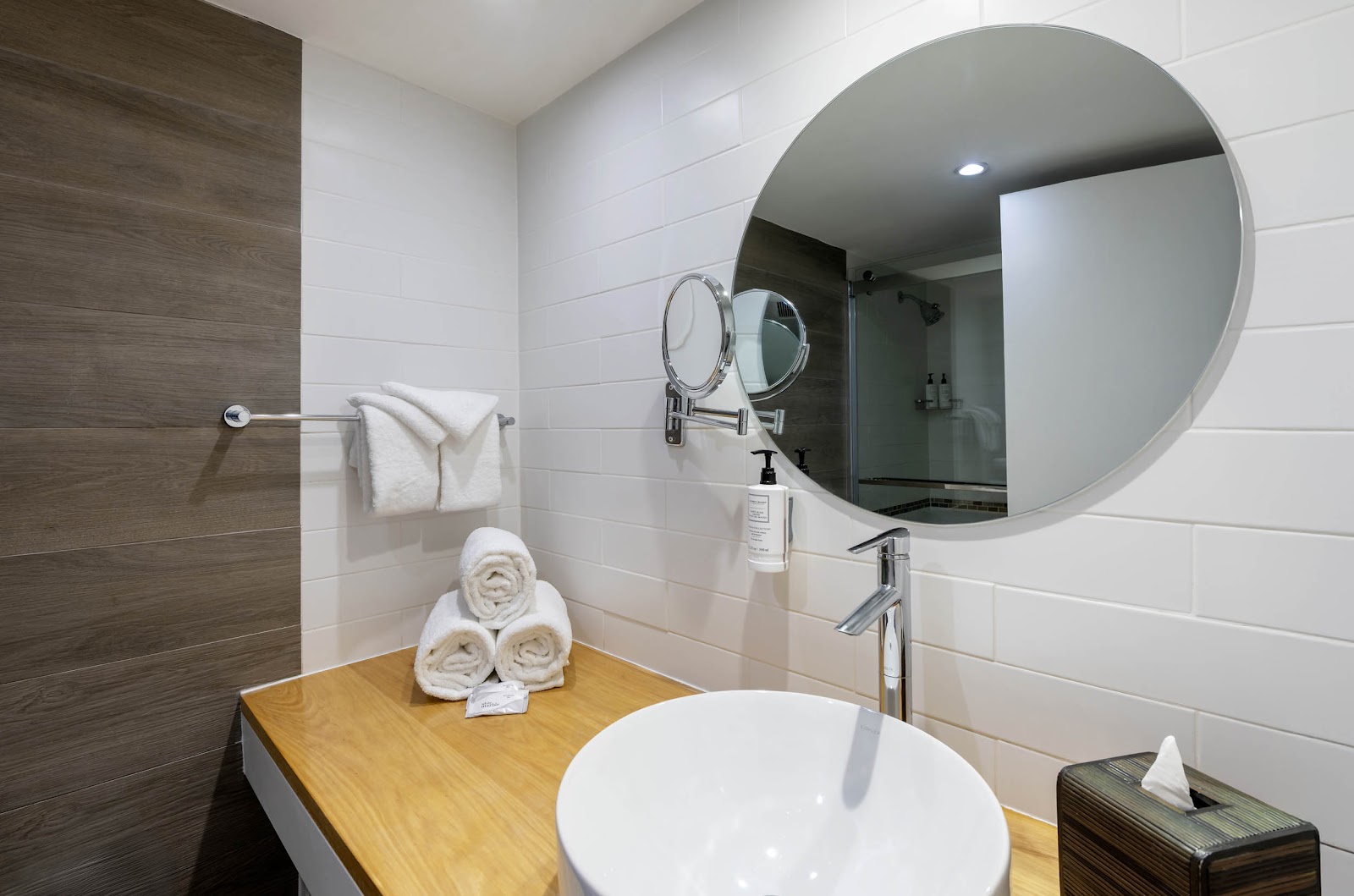 Modern bathroom with round mirror, white vessel sink, rolled towels on wooden counter, and dark wood accents.