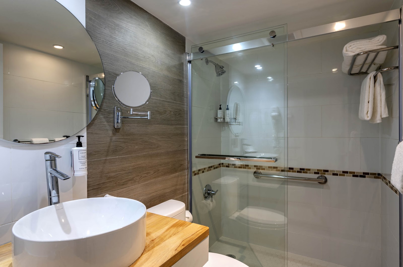Modern bathroom with round white vessel sink, wood-grain accent wall, circular mirror, and glass-enclosed shower with grab bars and towel rack.