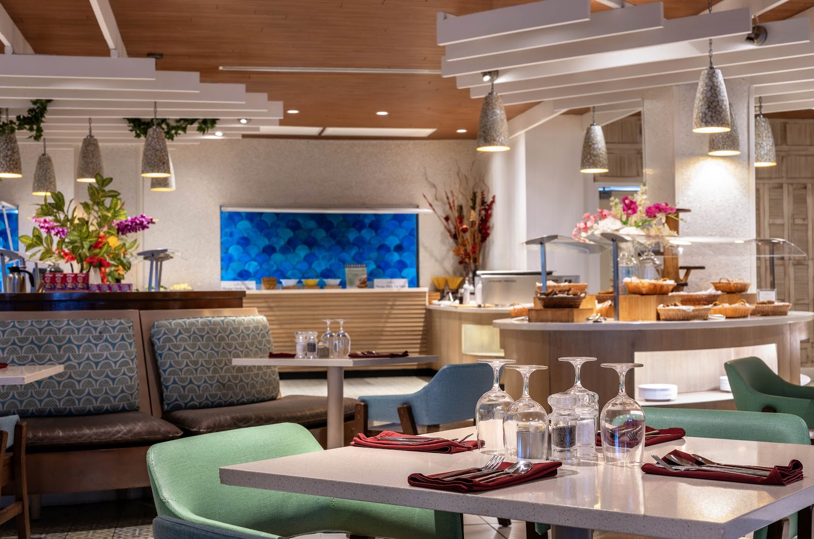 Modern resort dining area with green chairs, white tables set with glasses, wooden ceiling beams, pendant lights, and blue aquarium behind the reception desk.