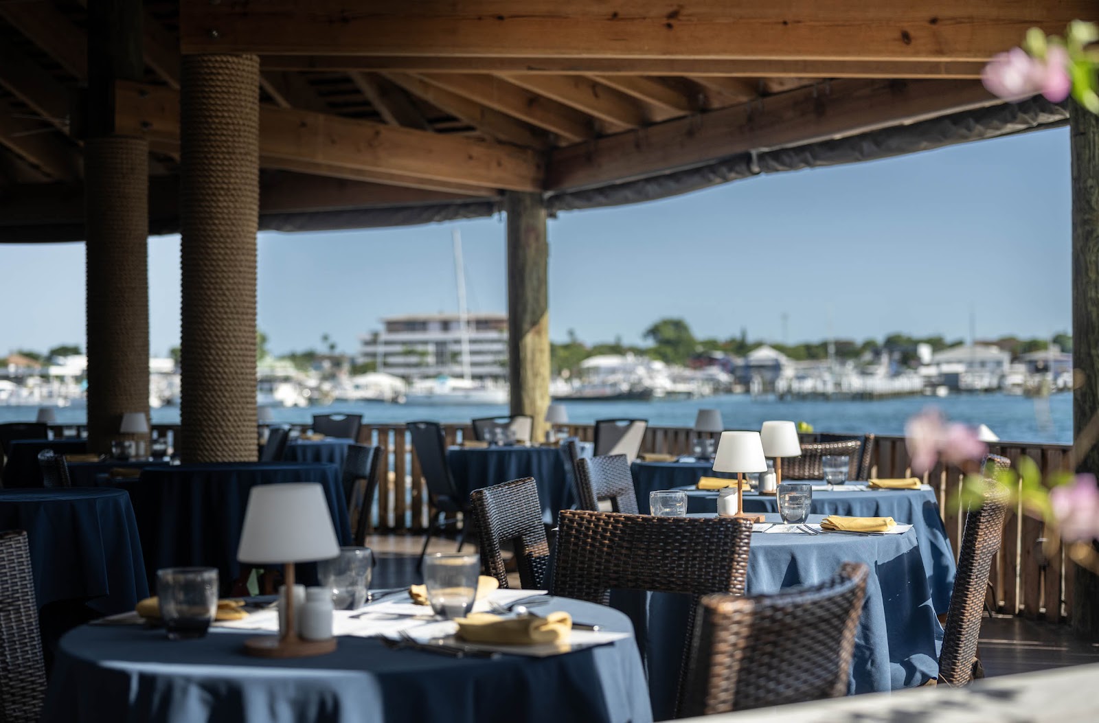 Waterfront dining pavilion with blue-clothed tables, rattan chairs, and a view of the harbor and coastal buildings beyond.