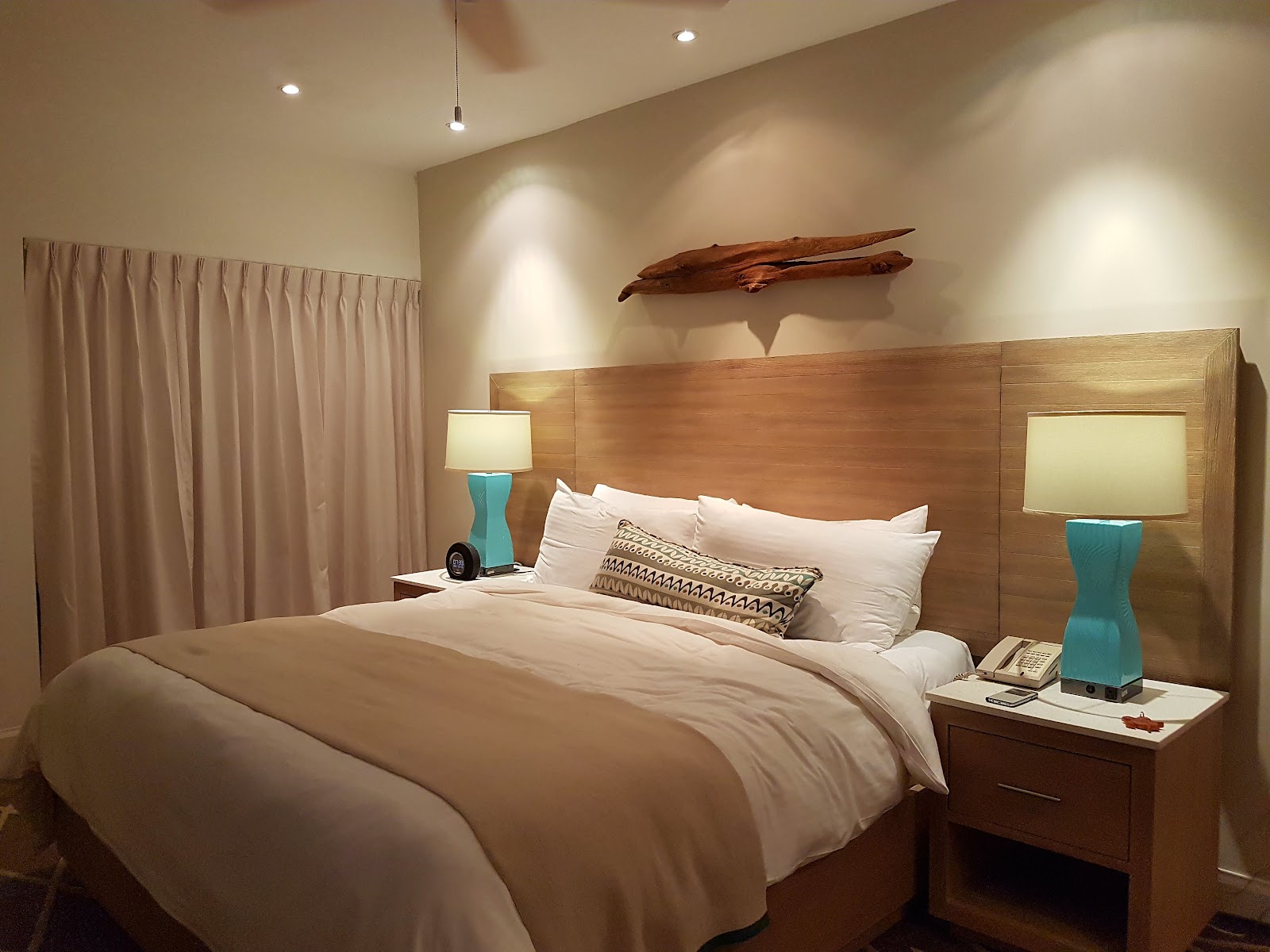 Resort bedroom with wood headboard, white bedding, turquoise lamps on nightstands, and driftwood wall art above the bed.