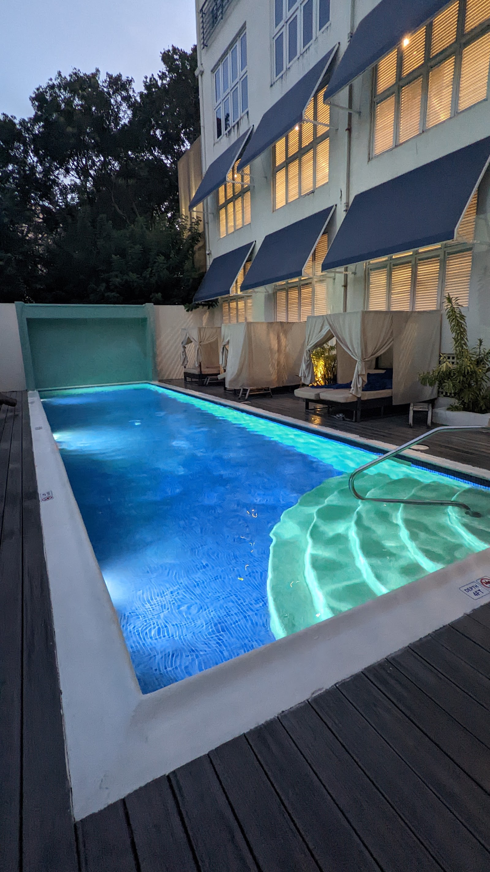 Illuminated pools on a wooden deck beside a white building with blue awnings and draped cabanas at dusk.