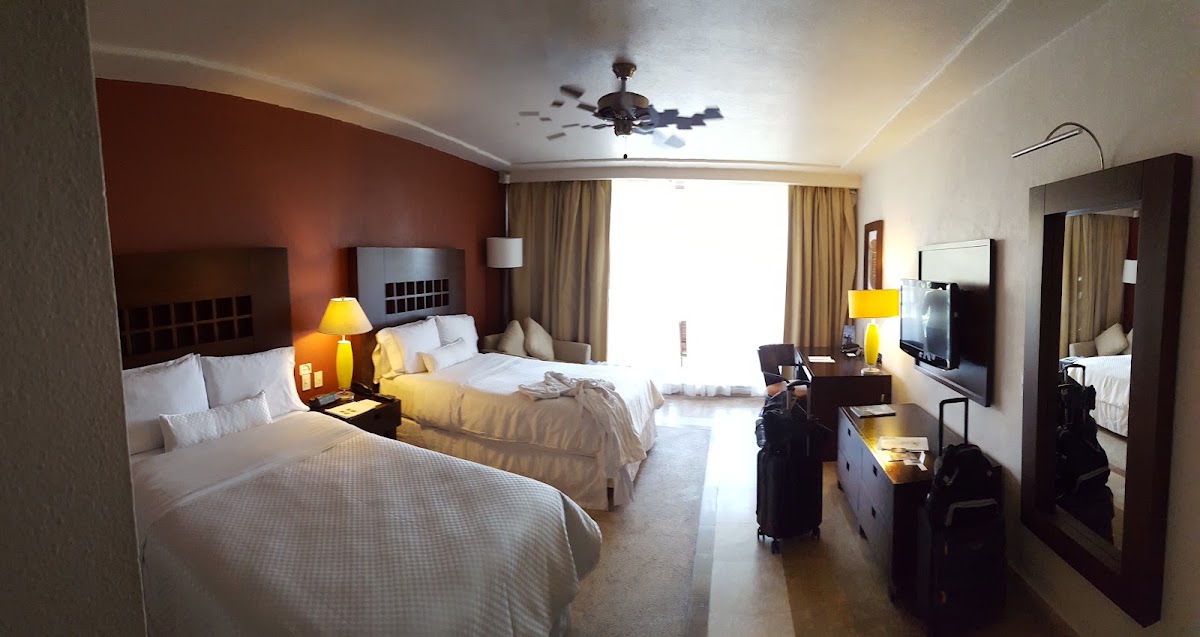 Double beds with white linens in a resort room with rust accent wall, dark wood furnishings, and balcony overlooking the ocean.