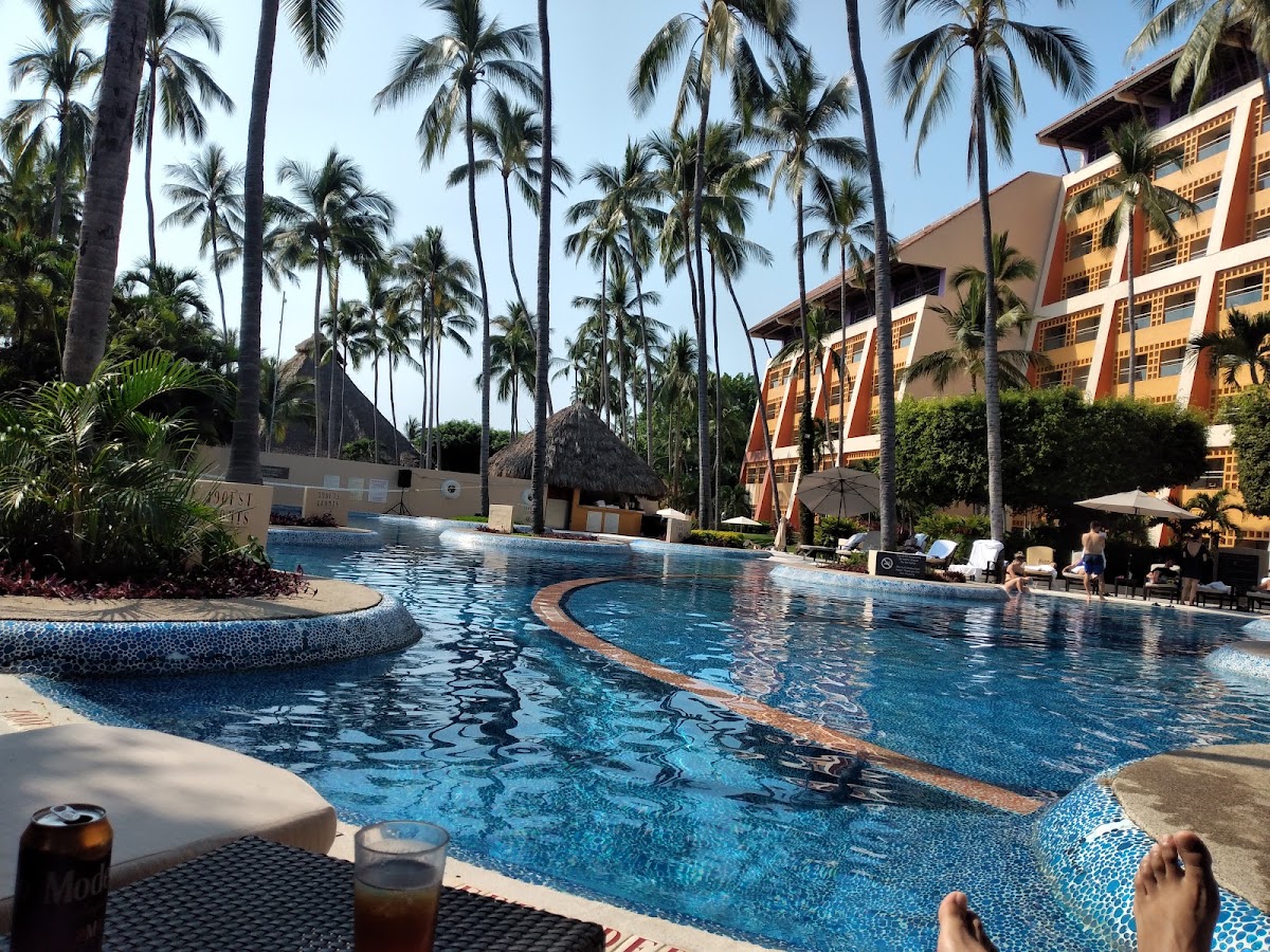 Multi-level pool with curved lanes surrounded by tall palm trees, thatched-roof bar, orange resort buildings, and guests relaxing on lounge chairs under umbrellas.