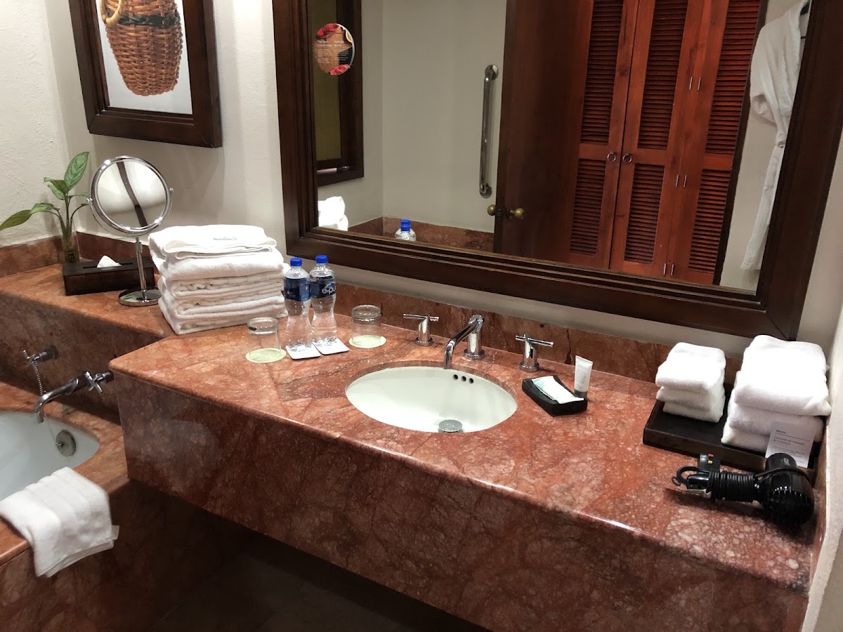 Resort bathroom with granite countertop, round white sink, chrome fixtures, stacked white towels, water bottles, and large mirror with wood frame.