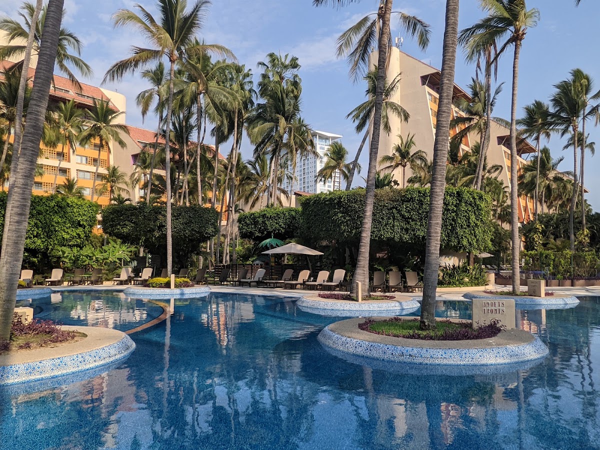 Resort pool with curved sections and mosaic tile trim surrounded by tall palm trees, lounge chairs, and tan multi-story buildings under clear sky.