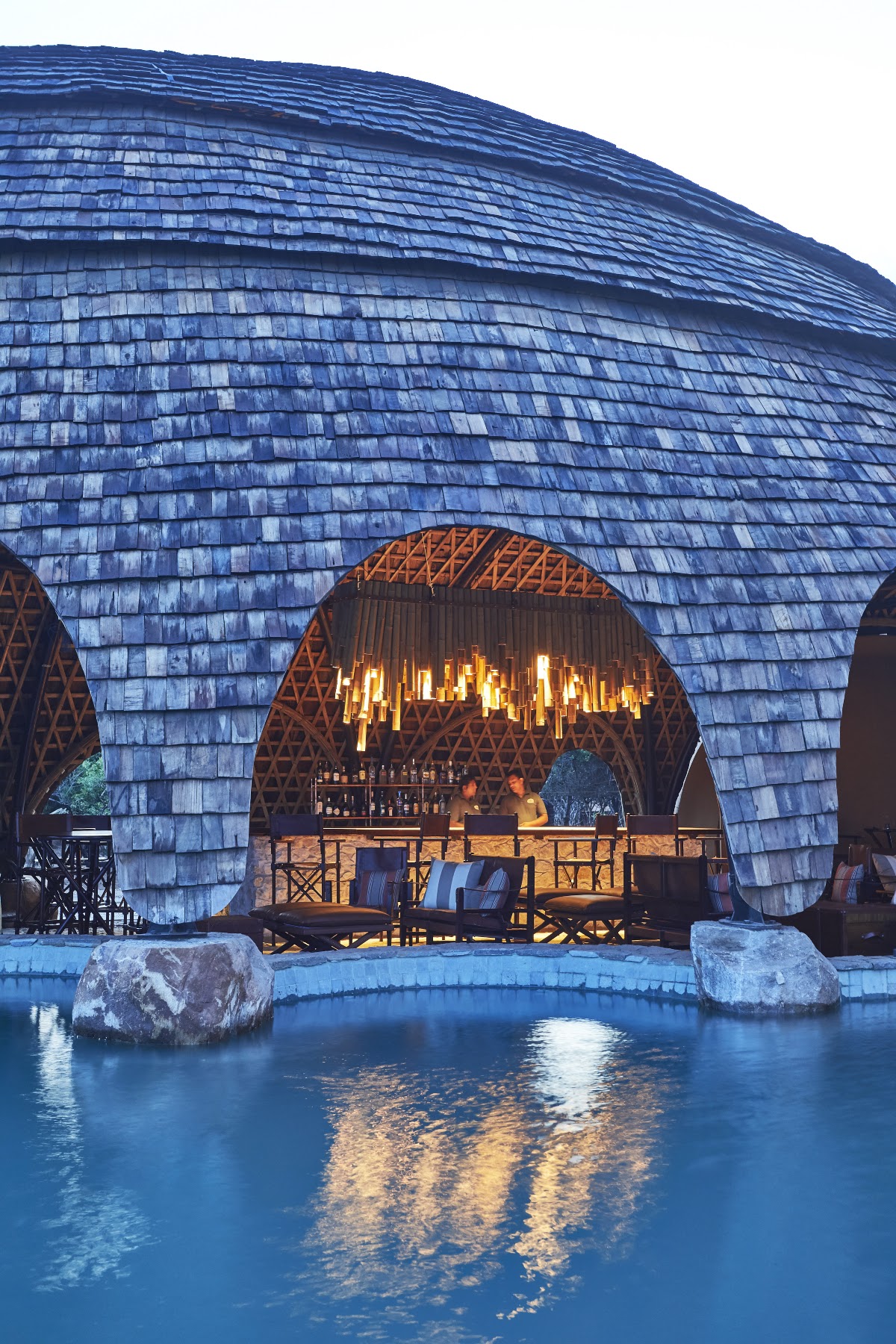 Blue slate-roofed dome structure at Wild Coast Tented Lodge with lit bar, wooden deck, and reflecting pool at dusk.
