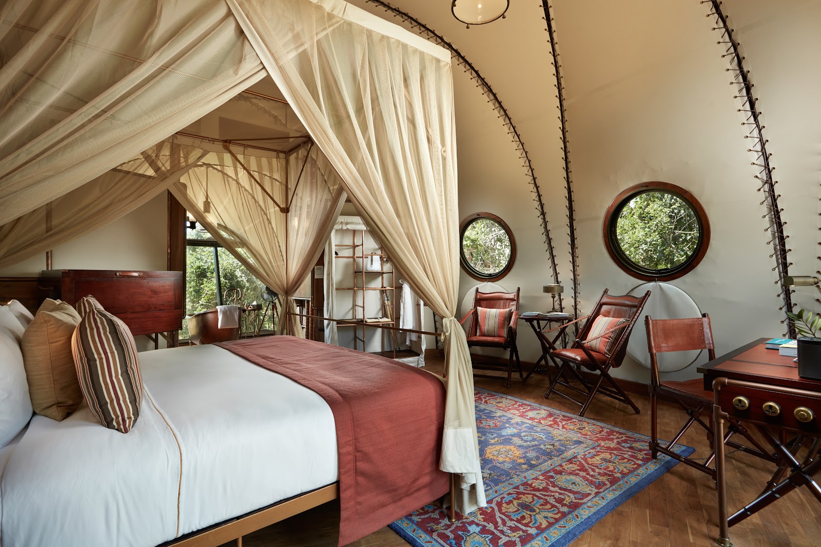Canopy bed with cream drapes and rust-colored runner in a tent-style room with circular windows, wooden furniture, and arched cactus branches overhead.