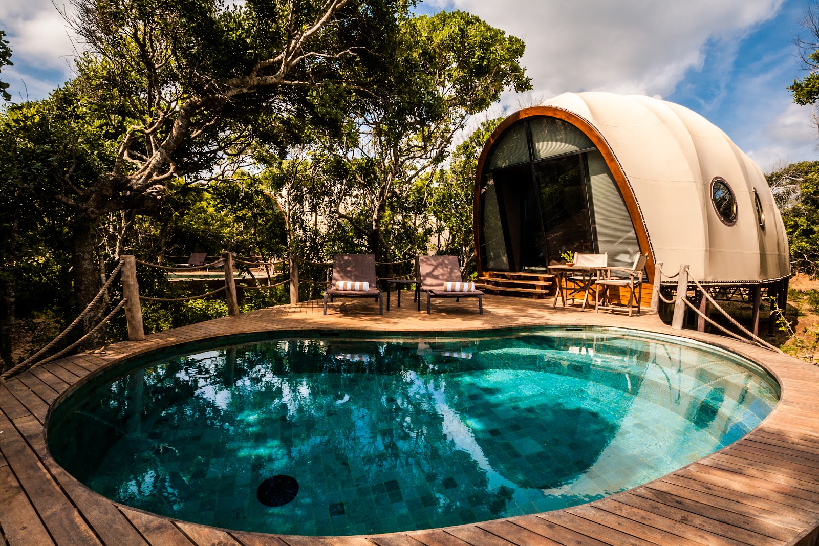 White glamping pod with orange trim beside a turquoise pool on a wooden deck, surrounded by tall trees and lounge chairs.