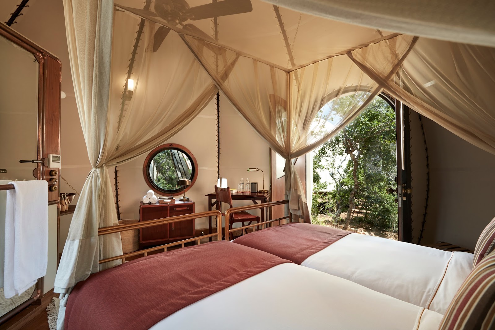 Glamping bedroom at Wild Coast Tented Lodge with draped canvas ceiling, twin beds, wooden vanity, and garden views through open doors.