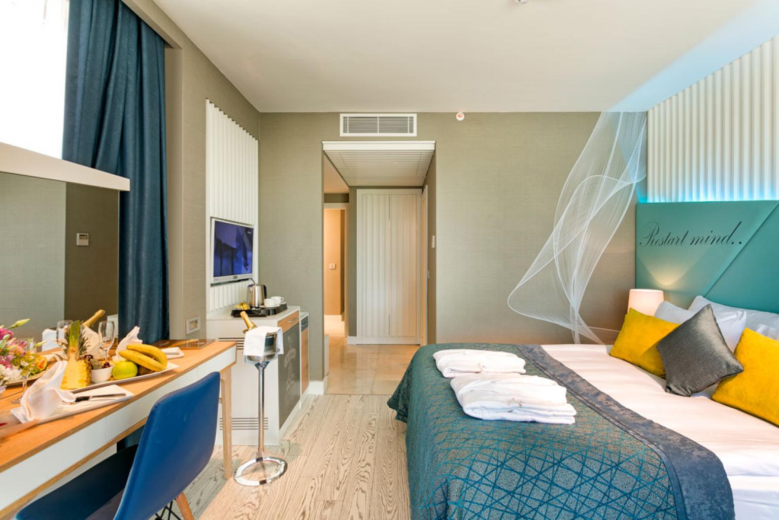 Bed with turquoise headboard featuring yellow and gray pillows, white sheer canopy, and bedside lamp in a modern resort room.