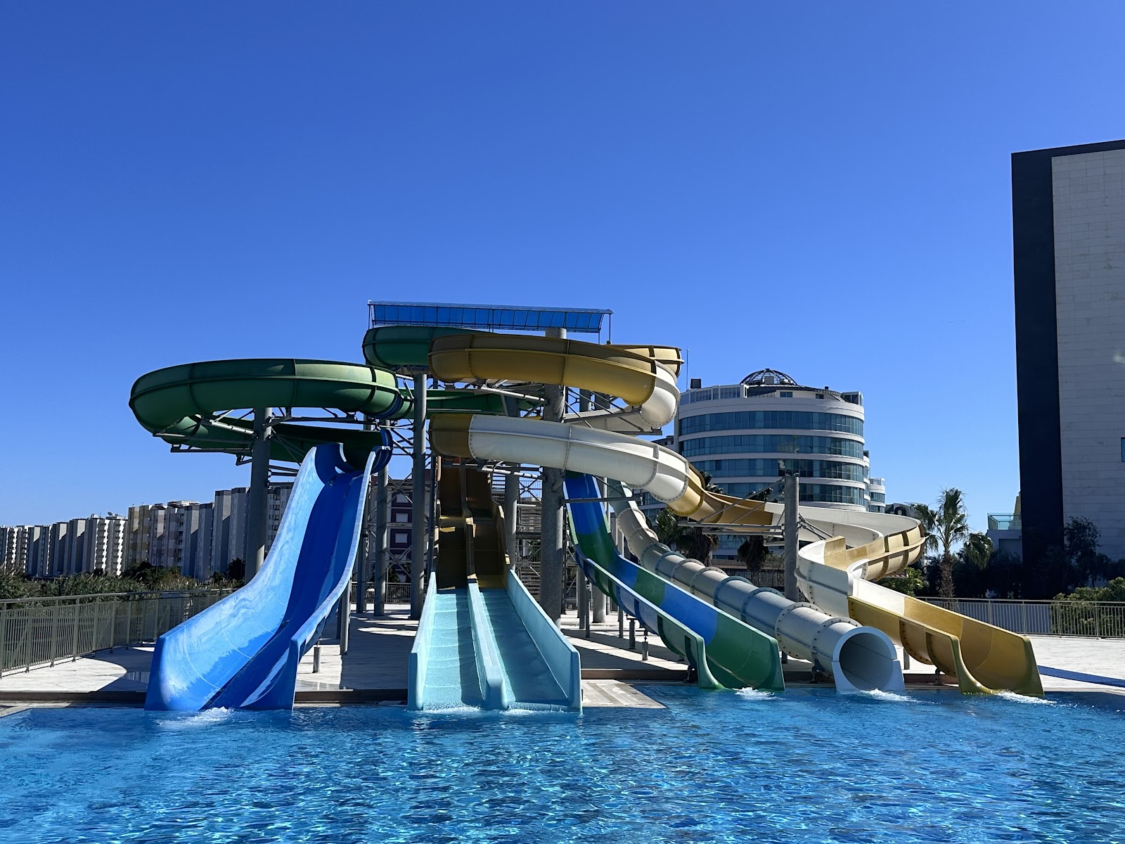 Water slides in blue, green, and yellow spiral into a pool at Wind of Lara Hotel & Spa, with resort buildings visible behind under clear sky.