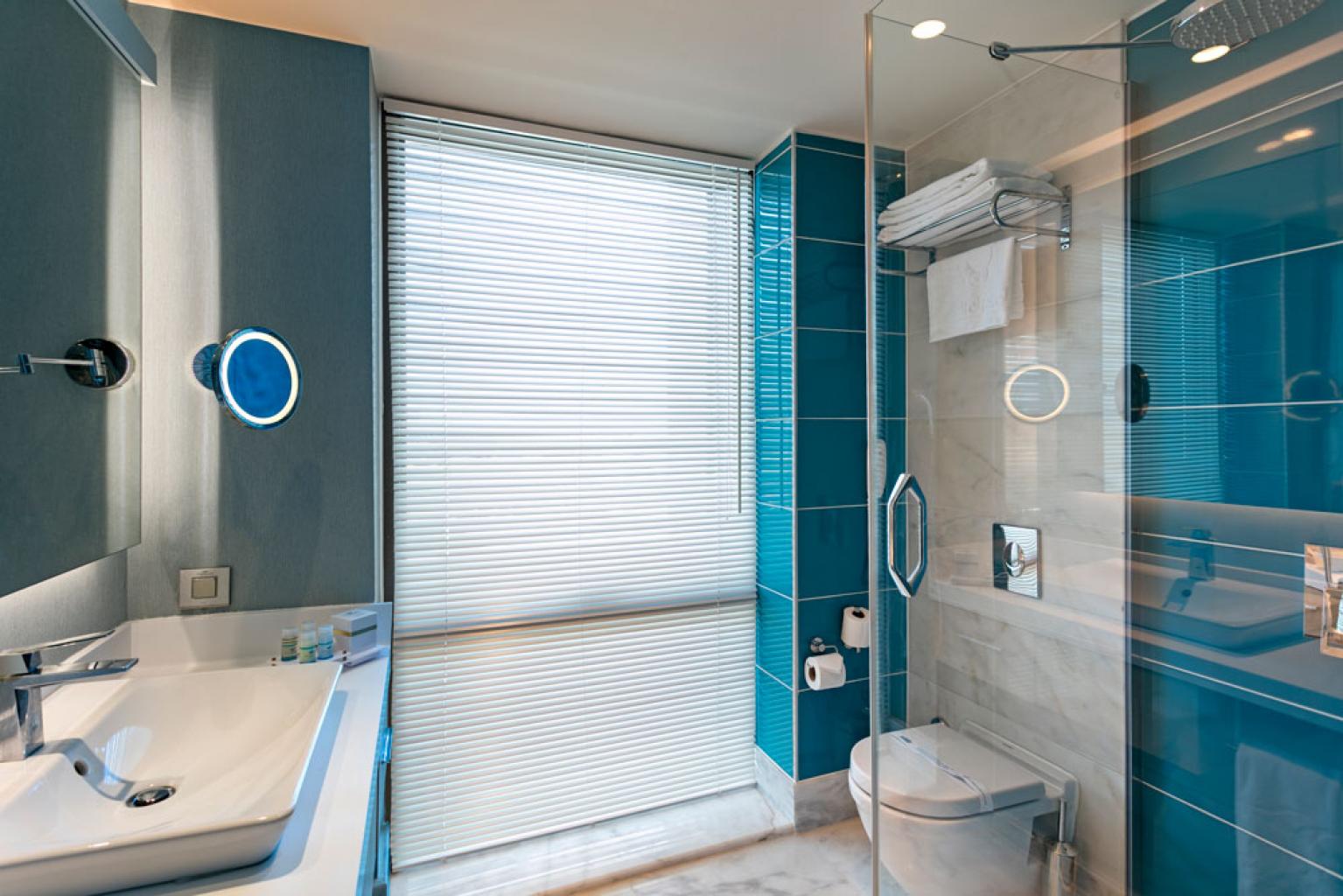 Modern bathroom with teal tile walls, glass shower enclosure, white sink, and round illuminated mirrors.