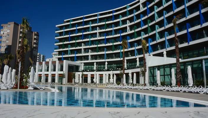 Wind of Lara Hotel & Spa's curved modern building rises behind a large pool surrounded by white loungers and palm trees under clear blue sky.