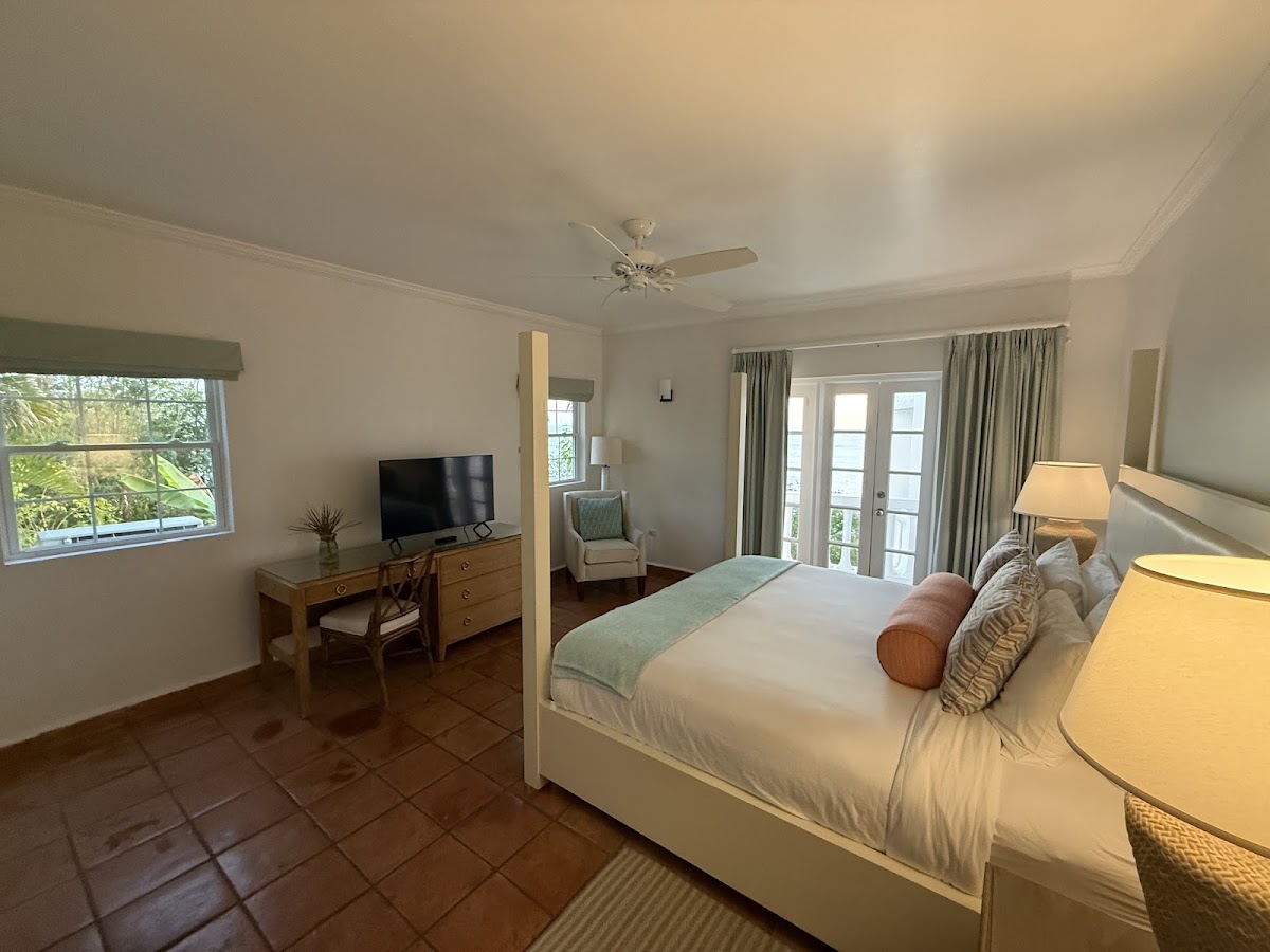 Spacious resort bedroom with king bed, TV, desk, sitting area, and balcony doors overlooking tropical grounds at Windjammer Landing.