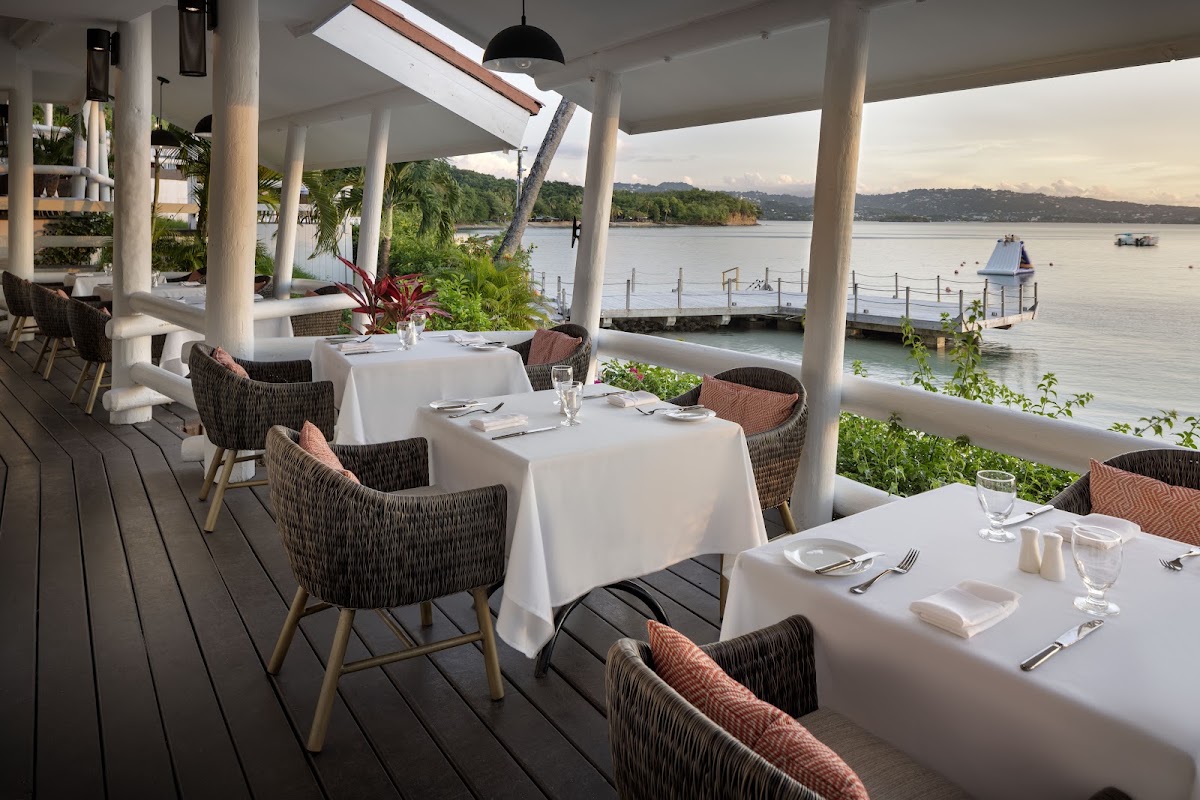Waterfront dining pavilion at Windjammer Landing with white-clothed tables, woven chairs, and views of the bay and forested coastline.