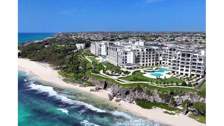 White multi-story resort buildings overlook a sandy beach and turquoise ocean with breaking waves at Wyndham Grand Barbados Sam Lords Castle.