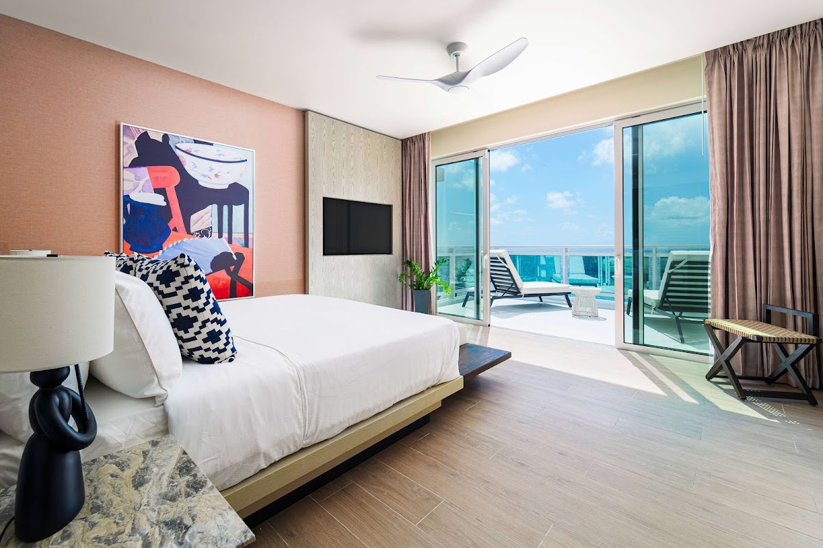 Modern resort bedroom with white bed, pink accent wall, and floor-to-ceiling ocean view windows opening onto a balcony with lounge chairs.