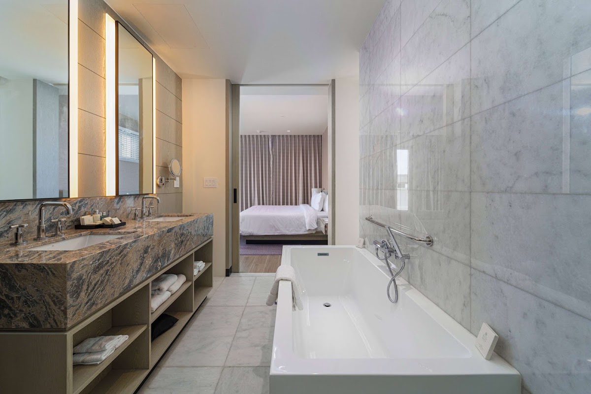 Marble bathroom with soaking tub, dual vanity with illuminated mirror, and view into bedroom with white bedding at Wyndham Grand Barbados Sam Lords Castle.