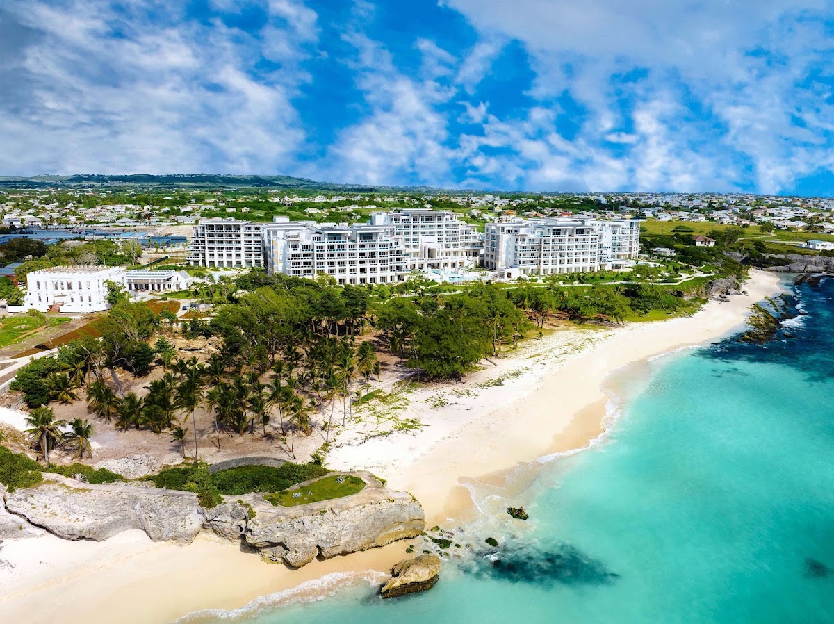 Modern multi-story resort buildings overlook a sandy beach with turquoise water, palm trees, and rocky outcrops along the Barbados coastline.